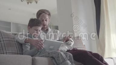 父亲和孩子用数字平板电脑坐在大客厅的沙发上。 爸爸教他的孩子。 快乐的爸爸和爸爸